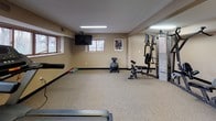This fitness center offers a well-rounded exercise environment housed within a single-level space with a practical, no-frills design. The room features durable speckled carpet flooring throughout, neutral beige walls, and flush-mounted fluorescent lighting that ensures consistent illumination across the entire area. A row of treadmills lines one side of the space, complemented by additional cardio equipment including a recumbent bike and elliptical machines, providing a variety of aerobic workout options. A multi-station cable weight machine serves as the centerpiece for strength training, accompanied by a weight bench and leg press attachment, offering users a comprehensive resistance workout. An open floor area is available for stretching and flexibility exercises, with a stability ball, resistance mat, and foam roller on hand. Wall-mounted flat-screen televisions are positioned for easy viewing from cardio equipment. Small windows along one wall allow modest natural light into the space. A desk and seating area provide a convenient spot for rest or planning. Framed artwork on the walls adds a mild decorative touch to the otherwise functional environment.