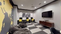 The Tech Lounge is a thoughtfully designed single-floor space that blends functionality with contemporary style. The room features a built-in workstation area along one wall, equipped with multiple desktop computers and vibrant yellow ergonomic chairs that add a bold pop of color against the neutral palette. Pendant lighting hangs above the desk surface, providing focused task illumination complemented by recessed ceiling lights throughout.

A striking black-and-white hashtag artwork serves as a visual focal point above the workstation, reinforcing the tech-forward identity of the space. On the opposite wall, a large flat-screen television is mounted at an ideal viewing height, supported by a warm wood-toned media console that adds contrast to the otherwise modern aesthetic.

The flooring features a distinctive checkered pattern in alternating light and dark carpet tiles, lending the space a dynamic, graphic quality. Plush, oversized faux-fur lounge seating in the foreground invites casual relaxation, creating a comfortable balance between a productive workspace and a social gathering area. Crown molding with dark trim accents adds a refined architectural detail to the room's overall design.
