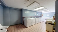 Located at 294 Main St in Hartford, Connecticut, this shared laundry facility offers a clean, functional, and well-organized space designed for efficient use. The room is finished with light blue painted walls and durable terracotta-toned ceramic tile flooring throughout, creating a practical and easy-to-maintain environment.

The main area is lined on both sides with rows of white top-load washing machines and front-load dryers, providing ample capacity for multiple users simultaneously. Each machine features a digital control panel, and usage instructions are posted on the walls for resident convenience. A continuous red-trimmed folding counter runs along one wall, offering a dedicated surface for sorting and folding laundry.

Overhead, recessed fluorescent panel lighting ensures the space is bright and evenly illuminated throughout. A dedicated waiting area near the entrance includes a row of modern white molded chairs, allowing residents to wait comfortably. A bulletin board displaying notices and informational signage is mounted on the wall nearby.

The space is accessed through wood-panel doors and is equipped with a fire extinguisher, smoke detectors, and a security camera, reflecting a strong emphasis on safety. Overall, the facility is straightforward in design and well-equipped to serve the practical laundry needs of its users.