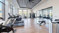 This fitness center at 12100 N Mountain Centre Rd in Marana, Arizona, offers a thoughtfully designed exercise space that balances functionality with modern aesthetics. The room is anchored by a distinctive raised tray ceiling adorned with a bold black-and-white geometric pattern and fitted with a large ceiling fan, creating a visually striking focal point overhead. Light wood-look flooring runs throughout, lending warmth to the space while remaining practical for high-traffic use.

Natural light pours in through a series of large, floor-to-ceiling windows along the exterior walls, providing open views of the surrounding Marana landscape and keeping the atmosphere bright and energizing. A full-length mirror along one wall further enhances the sense of openness and allows for easy form monitoring during workouts.

The facility is outfitted with a comprehensive selection of equipment, including multiple TRUE-brand treadmills and elliptical machines for cardio training, a multi-station cable and strength machine, an adjustable weight bench, and a rack of dumbbells covering a range of weights. Additional accessories such as stability balls and medicine balls are neatly organized on a dedicated storage rack near the entrance. The space is accessed through sleek, dark-framed double glass doors, reinforcing the clean, contemporary design that defines this well-appointed fitness facility.