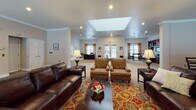 Located at 2900 Woods Edge Dr in Painted Post, NY, this single-floor facility offers a thoughtfully designed communal environment with a variety of functional and inviting spaces. The centerpiece of the property is a spacious common living area bathed in natural light from a large overhead skylight and recessed lighting. This room features plush seating arrangements including leather sofas and patterned armchairs set atop a colorful area rug, alongside a striking floor-to-ceiling stone fireplace that serves as a dramatic focal point. Large windows dressed with neutral drapes allow abundant natural light to fill the room, while French doors provide additional access and openness. Adjacent to the living area is a well-appointed dining nook furnished with a wooden table and wall-mounted television, ideal for casual gatherings. The fully equipped kitchen showcases rich wood cabinetry, stainless steel appliances including a range and refrigerator, granite countertops, and tile flooring with warm yellow accent walls. A reception area near the entrance features striped wallcovering, carpet flooring, a dark wood reception desk, and elegant glass-paned French doors leading to interior office spaces. Throughout the facility, crown molding, neutral tones, and carpeted floors contribute to a warm, welcoming, and professionally maintained atmosphere.