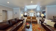 Located at 2900 Woods Edge Dr in Painted Post, NY, this single-floor facility offers a thoughtfully designed communal environment with a variety of functional and inviting spaces. The centerpiece of the property is a spacious common living area bathed in natural light from a large overhead skylight and recessed lighting. This room features plush seating arrangements including leather sofas and patterned armchairs set atop a colorful area rug, alongside a striking floor-to-ceiling stone fireplace that serves as a dramatic focal point. Large windows dressed with neutral drapes allow abundant natural light to fill the room, while French doors provide additional access and openness. Adjacent to the living area is a well-appointed dining nook furnished with a wooden table and wall-mounted television, ideal for casual gatherings. The fully equipped kitchen showcases rich wood cabinetry, stainless steel appliances including a range and refrigerator, granite countertops, and tile flooring with warm yellow accent walls. A reception area near the entrance features striped wallcovering, carpet flooring, a dark wood reception desk, and elegant glass-paned French doors leading to interior office spaces. Throughout the facility, crown molding, neutral tones, and carpeted floors contribute to a warm, welcoming, and professionally maintained atmosphere.