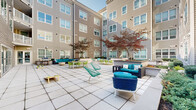Located at 198 College St in New Haven, Connecticut, this expansive outdoor courtyard serves as a central amenity space enclosed on multiple sides by a contemporary multi-story building clad in warm taupe horizontal siding with large, white-framed windows. The courtyard features a generously sized, large-format stone tile patio that provides a clean, modern foundation for the various activity zones throughout the space. Ornamental Japanese maple trees with rich burgundy and green foliage are planted throughout raised planters, adding seasonal color and a natural focal point to the hardscaped environment. Multiple distinct seating areas are thoughtfully arranged across the courtyard, including a large sectional sofa with teal cushions paired with a fire bowl, bistro-style dining tables with vibrant lime-green lattice chairs, and intimate lounge groupings featuring woven blue accent chairs and navy sofas with decorative pillows. A raised outdoor kitchen or bar structure anchors one section of the patio, while cornhole boards provide casual recreational entertainment. Large sliding glass and French doors connect the courtyard directly to interior common spaces, encouraging seamless indoor-outdoor flow. The surrounding building rises several stories, offering upper-level balconies that overlook the courtyard below, creating an engaging and community-centered outdoor living environment.