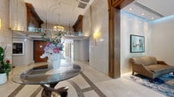 Located at 1100 Avenue du Docteur-Penfield in Montréal, this distinguished lobby combines classical architectural grandeur with refined contemporary touches. The entrance is anchored by a striking circular glass-top table with a sculptural twisted base, topped with a fresh floral arrangement, set atop an intricate mosaic-inlaid travertine tile floor. Soaring ceilings adorned with detailed plasterwork moldings and an upper-level mezzanine balcony lend the space a sense of impressive verticality. Warm wall sconces and a polished chandelier provide ambient lighting that complements the floor-to-ceiling travertine stone wall cladding and rich dark wood millwork framing the entryways.

The hallway leading further into the building is clean and well-lit, with elevator access and an administration office clearly marked, ensuring smooth circulation throughout. The adjacent lounge area serves as a welcoming reception space, featuring plush brown armchairs arranged on a vivid blue abstract area rug. Large floor-to-ceiling windows flood this room with natural light and offer serene views of the surrounding mature tree canopy. A framed antique map of Montréal serves as a focal artwork, reinforcing the building's connection to its storied urban setting. A wall-mounted display screen provides residents with building information, completing a lobby that is both functional and elegantly appointed.