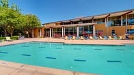 Located at 1580 Jade St in Davis, California, this outdoor recreational space is centered around a generously sized swimming pool complex that serves as the focal point of the community. The pool area features a large, freeform pool with clear turquoise water, complemented by an accessible pool lift, ensuring the space is welcoming to all users. A second, distinctly shaped pool is also present, offering additional aquatic options for residents and guests.

The surrounding pool deck is finished in warm-toned concrete, providing ample space for relaxation and socializing. Colorful lounge chairs and chaise lounges in blue and green are arranged throughout the deck and along the grassy perimeter, offering both sunny and shaded spots beneath a canopy of mature trees. The lush, well-maintained landscaping — featuring a mix of deciduous trees, manicured hedges, and patches of green lawn — creates a park-like atmosphere that softens the hardscape and provides natural shade.

The adjacent clubhouse building features expansive floor-to-ceiling glass walls and a covered overhang, allowing natural light to flood into the interior while maintaining a seamless visual connection to the outdoor pool area. The surrounding multi-story residential buildings add an urban context, while the open sky and mature tree cover contribute to a vibrant, resort-style ambiance suited to California's sunny climate.