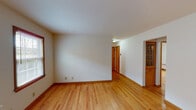 This single-floor space presents a clean, straightforward layout with a cohesive design aesthetic carried throughout. Hardwood flooring in a warm honey tone runs continuously across the living areas and bedrooms, complemented by matching wood baseboards and window trim that lend a consistent, classic character to the interior.

The living area is open and well-proportioned, with neutral white walls and access to additional rooms via wood-trimmed doorways. Multiple rooms feature windows fitted with horizontal blinds, allowing for controlled natural light. Several rooms are equipped with wall-mounted radiator units, providing an efficient and unobtrusive heating solution. A built-in glass-front cabinet adds a traditional touch in one area of the space.

The kitchen is practical and efficiently arranged, featuring warm wood cabinetry, a white refrigerator, a white stove, light countertops, and tile flooring. A ceiling fan provides additional ventilation, and a window over the counter brings in natural light.

The bathroom retains a retro charm with distinctive salmon-pink wall tile surrounding the tub-shower combination, mosaic tile flooring in muted tones, a white vanity, and a small frosted window. Overall, the space offers a functional and inviting atmosphere with consistent warm-toned finishes throughout.