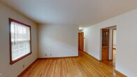This single-floor space presents a clean, straightforward layout with a cohesive design aesthetic carried throughout. Hardwood flooring in a warm honey tone runs continuously across the living areas and bedrooms, complemented by matching wood baseboards and window trim that lend a consistent, classic character to the interior.

The living area is open and well-proportioned, with neutral white walls and access to additional rooms via wood-trimmed doorways. Multiple rooms feature windows fitted with horizontal blinds, allowing for controlled natural light. Several rooms are equipped with wall-mounted radiator units, providing an efficient and unobtrusive heating solution. A built-in glass-front cabinet adds a traditional touch in one area of the space.

The kitchen is practical and efficiently arranged, featuring warm wood cabinetry, a white refrigerator, a white stove, light countertops, and tile flooring. A ceiling fan provides additional ventilation, and a window over the counter brings in natural light.

The bathroom retains a retro charm with distinctive salmon-pink wall tile surrounding the tub-shower combination, mosaic tile flooring in muted tones, a white vanity, and a small frosted window. Overall, the space offers a functional and inviting atmosphere with consistent warm-toned finishes throughout.