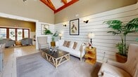 This thoughtfully designed single-floor space exudes coastal charm through a seamless blend of architectural character and refined interior details. The most striking feature upon entry is the dramatic vaulted ceiling adorned with bold, exposed natural wood beams that draw the eye upward, amplifying the sense of openness throughout the living area. White shiplap walling runs along the lower half of the walls, lending a clean, beachy aesthetic that is complemented by warm taupe tones above.

The entrance area is welcoming and spacious, featuring stone-tile flooring, a built-in bench with accent pillows, and oversized French doors with leaded glass that flood the space with natural light. Multiple seating arrangements — including plush upholstered armchairs and a neutral sofa — create comfortable gathering zones anchored by patterned area rugs.

The kitchen is well-equipped and stylish, showcasing floor-to-ceiling white cabinetry, dark stone countertops, stainless steel appliances, and a central island with wicker bar seating. Large windows continue to bring in generous natural light, maintaining the airy coastal atmosphere. Decorative botanical bird artwork, wall sconces, and lush potted plants add warmth and personality throughout, completing this cohesive and inviting space.