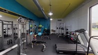 This single-floor fitness facility is designed to support a comprehensive workout experience, combining cardio and strength-training equipment within a compact yet efficiently organized space. The interior is distinguished by its bold color scheme, featuring teal accent walls paired with a yellow-green ceiling, which give the space a vibrant, energetic atmosphere. Exposed ductwork runs along the ceiling, adding an industrial character, while pendant lighting provides adequate illumination throughout.

The main workout area is well-stocked with a variety of equipment, including multiple Precor treadmills, an elliptical machine, and a cable weight machine. A dedicated free weights section offers a range of dumbbells, kettlebells, and medicine balls neatly arranged on racks. Additional strength equipment includes a squat rack with weight plates, a flat bench, and an adjustable bench. Stability balls in multiple sizes are also available for functional training exercises.

A large window along one wall allows natural light to fill the cardio zone, providing views of the surrounding outdoor area. Rubber flooring covers the entire workout space, offering durability and safety underfoot. A utility area near the entrance includes dual wall-mounted water fountains and access to a restroom, ensuring user convenience throughout a workout session.