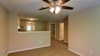This single-floor residence offers a practical and comfortable layout designed for everyday living. The main living area is finished with wall-to-wall carpeting in a neutral tone, creating a warm and inviting atmosphere. A five-blade ceiling fan with an integrated light fixture is centrally mounted, providing both airflow and ambient illumination throughout the space.

A half-wall partition separates the living area from the kitchen, offering an open-concept feel while maintaining a degree of definition between the two spaces. The kitchen area, visible beyond the partition, features stainless steel appliances and benefits from natural light entering through a window. Flush-mount ceiling lighting in the kitchen ensures consistent brightness.

Neutral beige wall tones are carried throughout the interior, contributing to a cohesive and understated aesthetic. White trim and baseboards provide clean contrast along the walls and doorways. A central hallway leads from the living area to additional rooms, with white-paneled doors offering access to each space. The overall layout is straightforward and functional, making efficient use of the available floor plan. The textured ceiling adds a subtle traditional character to the interior throughout.