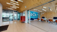 Located in the South Congress neighborhood of Austin, Texas, this single-floor space is a standout example of contemporary design. The interior is immediately distinguished by its angled natural pine wood ceiling, which runs throughout the main corridor and common areas, lending warmth and architectural character to the space. Large-format tile flooring and expansive floor-to-ceiling glass walls create a bright, open atmosphere, allowing natural light to flow freely throughout.

The leasing and reception area greets visitors with a bold "SoCo"-branded accent wall rendered in vivid blue tilework, paired with a modern workstation and stylish seating in navy and orange tones. Adjacent to the entrance, a welcoming lounge area features a striking high-back red accent chair and curated wall art, adding a residential warmth to the professional setting.

A standout lounge space showcases a dramatic navy blue upholstered banquette with vibrant orange accent pillows, complemented by red armchairs, round side tables, and a soft area rug — creating an inviting gathering area. Glass-partitioned rooms line the corridor, offering views of additional meeting and collaboration spaces with distinctive tiled feature walls. Abundant glazing throughout provides continuous sightlines to the surrounding greenery and outdoor areas, reinforcing a seamless connection between interior and exterior.
