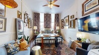 Located steps from San Antonio's historic Main Plaza, this compact yet characterful studio offers a single-floor layout comprising a living area, kitchen, bathroom, closet, and entrance. The main living space is richly decorated with an eclectic mix of framed artwork, ornate mirrors, and decorative collectibles that create a warm, gallery-like atmosphere. A wrought-iron bed anchors the room beneath a ceiling fan, while bold patterned drapes frame windows that invite natural light and offer views of the surrounding cityscape. A Persian-style area rug and a checkerboard accent rug layer texture underfoot, complementing the wood-tone flooring throughout. A wooden ladder bookshelf adds vertical storage and displays trailing houseplants, adding a lush, lived-in charm. The compact kitchen is efficiently arranged with light wood cabinetry, white appliances, a tile backsplash, and a countertop sink. A dedicated seating nook with an armchair and wall-mounted TV provides a distinct lounge area. The bathroom features classic white subway tile wainscoting, vintage-style green mosaic floor tile, and a freestanding towel rack, maintaining the space's timeless character. Every corner of this studio is carefully styled, making it a distinctive and inviting retreat in downtown San Antonio.