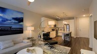 This single-floor residence showcases an open-concept design that unifies the living and kitchen areas into one cohesive, functional space. The living area is anchored by a ceiling fan overhead and features warm ambient lighting provided by a sleek arc floor lamp and a classic table lamp, creating an inviting atmosphere. Rich, dark hardwood flooring runs throughout, offering a striking contrast to the neutral, light-toned walls.

The kitchen is well-appointed with white cabinetry, stainless steel appliances including a refrigerator, range, and microwave, and a functional layout that maximizes efficiency. A kitchen island with pendant lighting above serves as both a prep surface and a casual dining spot, with bar-height seating adding to its versatility. A delicate chandelier above the island adds a refined decorative touch.

The living space comfortably accommodates seating, including a cushioned sofa and a stylish lounge chair with ottoman, arranged on a patterned area rug. Large artwork on the wall adds a bold visual element. The overall ambiance is contemporary and polished, balancing practicality with modern aesthetic appeal.