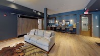 This single-floor communal space is thoughtfully designed to serve multiple functions while maintaining a cohesive, contemporary aesthetic throughout. Rich wide-plank hardwood flooring anchors the entire layout, complemented by a bold deep teal and neutral gray color palette that gives the space a modern yet inviting character.

The main lounge area is generously proportioned, featuring multiple plush neutral-toned sofas arranged around a striking live-edge wood coffee table set atop a textured area rug — a standout natural centerpiece that adds an organic, artistic touch. Accent pillows with geometric patterns and coordinating rugs further elevate the design.

Large windows along one wall flood a seating alcove with natural light, offering views to an outdoor area and providing an airy, open feel. This nook is furnished with upholstered armchairs and a warm wood credenza topped with table lamps and a decorative mirror.

A built-in banquette dining area lines one wall, with patterned upholstered seating and dark pedestal tables, illuminated by a row of sleek pendant lights overhead — ideal for casual gathering or dining.

Adjacent to the main lounge, a dedicated business center features multiple computer workstations set into a custom built-in desk beneath pendant lighting, creating a focused yet stylish workspace. Rustic barn-style sliding wood doors and exposed structural columns add architectural character throughout, tying together the industrial and refined elements of this well-appointed space.