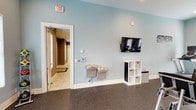 This fitness center is a comprehensive, single-floor facility designed to support a full range of workout routines. The space is finished with soft blue-gray walls and durable speckled rubber flooring throughout, creating a clean, energizing atmosphere conducive to exercise.

The main workout area is generously sized and hosts an array of cardio equipment, including treadmills and elliptical machines, each equipped with individual display screens. A large wall-mounted mirror along one side visually expands the space and allows users to monitor their form during workouts. Strength training is well-represented with a multi-station cable machine, free weights, and additional resistance equipment arranged for efficient use of the floor space.

A dedicated stretching and flexibility zone features wall-mounted yoga mat racks stocked with blue exercise mats, a stack of fitness balls, and a large floor mat, providing ample room for warm-ups, cool-downs, and mat-based exercises. Natural light flows in through multiple windows fitted with blinds, offering views of the surrounding greenery.

The entryway area includes dual wall-mounted water fountains for convenient hydration, a wall-mounted flat-screen television, a cubby storage unit, and a wall clock. The facility connects to an adjacent corridor, rounding out a practical and well-appointed fitness environment.