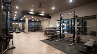 Located at 548 Newtown Rd in Virginia Beach, Virginia, this well-appointed fitness facility offers a thoughtfully designed single-floor layout built to support a wide range of workout routines. The main gym floor is expansive and open, finished with light wood-look flooring that contrasts cleanly against the dark ceiling and exposed black ductwork — an industrial design detail that adds a modern, urban character to the space. Structural columns are integrated seamlessly throughout, and recessed lighting provides bright, even illumination across the entire floor.

The strength training zone is well-equipped with a full rack of dumbbells, power racks, weight benches, and resistance machines, all positioned along mirrored walls that help amplify the sense of space. A dedicated cardio section houses treadmills, ellipticals, and stationary bikes, arranged to allow comfortable movement between machines. Accent walls in deep navy blue add visual depth and a professional atmosphere to the space.

A separate yoga studio is accessible through glass-paneled French doors, offering a quieter, dedicated environment for low-impact training and stretching. The studio features the same wood-look flooring as the main gym for a cohesive aesthetic throughout. Sanitation stations with equipment wipes are conveniently placed near studio entrances, reflecting a commitment to cleanliness and user experience.
