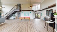 Located at 11502 Craig Ct in Saint Louis, MO, this single-floor community clubhouse offers a generously sized, open-concept layout designed for both relaxation and recreation. The space is defined by its dramatic double-height ceilings, which create an airy, expansive atmosphere throughout.

The living room area serves as the heart of the space, anchored by a sleek, modern fireplace set against a bold accent wall. A wall-mounted flat-screen television sits above the fireplace, while multiple seating arrangements — including plush sofas accented with teal throw pillows — invite comfortable gatherings. Exposed industrial-style ductwork adds a contemporary architectural detail that contrasts elegantly with the light hardwood flooring running throughout.

An abundance of large, multi-pane windows lines the walls on multiple sides, flooding the interior with natural light and offering views of the surrounding outdoor greenery. A staircase with a classic wooden railing leads to an upper landing, adding vertical dimension to the layout.

The recreational side of the space features a full-sized billiards table as a central attraction, complemented by a kitchenette bar with counter seating — ideal for social events. Ceiling fans with pendant lighting provide both comfort and style, while French doors open to the exterior, seamlessly connecting the indoor and outdoor environments.