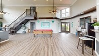Located at 11502 Craig Ct in Saint Louis, MO, this single-floor community clubhouse offers a generously sized, open-concept layout designed for both relaxation and recreation. The space is defined by its dramatic double-height ceilings, which create an airy, expansive atmosphere throughout.

The living room area serves as the heart of the space, anchored by a sleek, modern fireplace set against a bold accent wall. A wall-mounted flat-screen television sits above the fireplace, while multiple seating arrangements — including plush sofas accented with teal throw pillows — invite comfortable gatherings. Exposed industrial-style ductwork adds a contemporary architectural detail that contrasts elegantly with the light hardwood flooring running throughout.

An abundance of large, multi-pane windows lines the walls on multiple sides, flooding the interior with natural light and offering views of the surrounding outdoor greenery. A staircase with a classic wooden railing leads to an upper landing, adding vertical dimension to the layout.

The recreational side of the space features a full-sized billiards table as a central attraction, complemented by a kitchenette bar with counter seating — ideal for social events. Ceiling fans with pendant lighting provide both comfort and style, while French doors open to the exterior, seamlessly connecting the indoor and outdoor environments.
