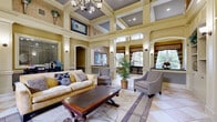 Located in Largo, Florida, this sophisticated single-floor space exudes a grand, clubhouse-style ambiance through its impressive architectural detailing and refined interior design. The living area commands attention with dramatically soaring coffered ceilings adorned with recessed lighting and an elegant chandelier, creating a sense of openness and grandeur. Cream-toned walls, arched doorways, and decorative columns frame the space beautifully, while large-scale tile flooring in a classic diamond pattern runs throughout, unifying the layout.

The living room is appointed with plush, neutral-toned seating, a richly stained wood coffee table, and an area rug that anchors the arrangement. Built-in shelving and arched alcoves add both function and visual interest, while lush indoor plants introduce a natural touch.

The dining area offers an intimate setting beneath high ceilings, featuring a round glass-topped table surrounded by dark wood chairs with upholstered seats. A textured accent wall with framed artwork and decorative drapery with striped valances adds warmth and character. Large windows flood the space with natural light and offer views of a shimmering outdoor pool, seamlessly connecting the interior to its Florida surroundings. A built-in counter with dark stone surfaces provides a practical serving or display area nearby.