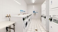 Located at 450 Rue Sherbrooke E in Montréal, this shared laundry facility offers a clean, well-organized space designed for efficient, high-volume use. The room is finished throughout with white ceramic floor tiles and white glazed wall tiles, creating a bright and sanitary environment that is easy to maintain. Overhead fluorescent lighting ensures the space is well-lit at all times.

The facility is equipped with a row of front-loading washing machines along one wall, complemented by multiple stacked dryer units along the opposite wall, maximizing the available floor space. A utility sink with brass fittings is conveniently positioned near the entrance, and a folding table provides a dedicated surface for sorting and handling laundry. A stainless steel waste bin is centrally placed for easy access.

Payment is handled through a modern card-based kiosk mounted on the wall, accompanied by posted instructional signage to guide users through the process. A wall-mounted split air conditioning unit ensures comfortable temperatures year-round. The room is accessed through a solid door with a glazed panel, and a floor drain is integrated into the tiled flooring for practical maintenance. Overall, the space is thoughtfully laid out to accommodate multiple users simultaneously while maintaining a tidy, functional atmosphere.