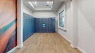 Located at 12100 N Mountain Centre Rd in Marana, Arizona, this single-floor package locker facility is designed to provide a convenient and secure parcel management solution. The space is divided into two connected rooms, each finished with warm wood-look luxury vinyl plank flooring and clean white walls with crisp white baseboard trim throughout.

The primary room is the focal point of the facility, housing an expansive bank of smart parcel lockers lining two walls in a corner configuration. The navy-blue locker units feature a centrally mounted digital touchscreen interface, allowing for seamless package retrieval. A tray ceiling detail adds a subtle architectural accent overhead, while a large picture window along one wall admits natural light and offers views of the surrounding residential community, including a glimpse of a pool and neighboring structures.

The secondary anteroom serves as an entry point to the space, accessible through a glass-paneled door that allows sightlines between the two areas. A narrow transom window above the door further enhances light flow. A vibrant wall mural adds a decorative touch, referencing the rich canyon landscapes characteristic of the greater Arizona region. The overall design is modern, functional, and well-lit, reflecting a purpose-built amenity space suited for a contemporary residential community.