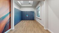 Located at 12100 N Mountain Centre Rd in Marana, Arizona, this single-floor package locker facility is designed to provide a convenient and secure parcel management solution. The space is divided into two connected rooms, each finished with warm wood-look luxury vinyl plank flooring and clean white walls with crisp white baseboard trim throughout.

The primary room is the focal point of the facility, housing an expansive bank of smart parcel lockers lining two walls in a corner configuration. The navy-blue locker units feature a centrally mounted digital touchscreen interface, allowing for seamless package retrieval. A tray ceiling detail adds a subtle architectural accent overhead, while a large picture window along one wall admits natural light and offers views of the surrounding residential community, including a glimpse of a pool and neighboring structures.

The secondary anteroom serves as an entry point to the space, accessible through a glass-paneled door that allows sightlines between the two areas. A narrow transom window above the door further enhances light flow. A vibrant wall mural adds a decorative touch, referencing the rich canyon landscapes characteristic of the greater Arizona region. The overall design is modern, functional, and well-lit, reflecting a purpose-built amenity space suited for a contemporary residential community.