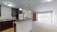 This single-floor unit is designed with an open-concept layout that seamlessly connects the kitchen and living area. The kitchen is well-appointed with rich, dark espresso cabinetry offering generous storage, light countertops along the perimeter, a granite-topped central island with pendant lighting overhead, and a full suite of stainless steel appliances including a range, built-in microwave, dishwasher, and refrigerator. Light hardwood-style flooring runs throughout the kitchen, transitioning to plush wall-to-wall carpeting in the living area.

The living space is bright and open, benefiting from vaulted ceilings that enhance the sense of volume, and a large sliding glass door fitted with vertical blinds that leads to an outdoor balcony and brings in natural light. Additional rooms include a carpeted bedroom with a dedicated window providing outdoor views, and a bathroom featuring a dark vanity with a white countertop, undermount sink, and large mirror. A walk-in closet with built-in shelving provides practical storage. Throughout the unit, warm wood-toned doors and trim create a cohesive, welcoming aesthetic.