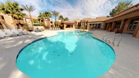This expansive outdoor amenity area delivers a true resort-style experience, centered around a generously sized, freeform swimming pool with crystal-clear turquoise water. The pool's curvilinear design creates an inviting, organic aesthetic, complemented by a separate bubbling spa that offers a relaxing hydrotherapy option. Decorative flower beds with vibrant seasonal blooms are thoughtfully integrated into the pool surround, adding color and charm to the overall landscape.

The surrounding pool deck is finished in a light-toned concrete surface, providing ample space for sunbathing and leisure. Multiple rows of chaise lounges are arranged along the deck, offering plentiful spots to relax and soak in the sun. Shaded dining areas with tables, chairs, and a bold green umbrella provide a comfortable retreat from direct sunlight.

A covered outdoor kitchen pavilion features a full-sized stainless steel gas grill, counter space, and seating — ideal for outdoor cooking and entertaining while enjoying direct views of the pool. The entire area is enclosed by a wrought iron fence and is framed by towering palm trees that lend a warm, desert-resort ambiance. The surrounding architecture features earth-toned stucco buildings with tile rooflines, consistent with a Southwestern design aesthetic.