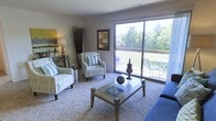 This single-floor living space offers a welcoming and well-appointed interior designed for both comfort and functionality. The living area is carpeted and bathed in natural light, thanks to a large sliding glass door that serves as a prominent architectural feature. This expansive glass opening not only floods the room with daylight but also provides direct access to a private balcony, where outdoor seating is visible against a backdrop of mature, verdant trees — creating a seamless indoor-outdoor connection.

The living room is thoughtfully arranged with patterned accent chairs, a glass-top coffee table, and a sofa, giving the space a casual yet coordinated aesthetic. Decorative elements, including tall dried branch arrangements, framed wall art, and tabletop accessories, contribute to the room's polished character. A console table along one wall adds both style and practical storage.

The neutral color palette of the walls and carpet provides a versatile backdrop that enhances the sense of openness throughout the space. Overall, the living area strikes a balance between relaxed comfort and tasteful design, with the balcony and surrounding greenery serving as a particularly notable highlight.