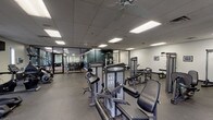 This single-floor fitness facility is thoughtfully organized into distinct training zones, each designed to support a comprehensive workout experience. The space features a drop-panel ceiling with recessed fluorescent lighting and ceiling fans throughout, ensuring consistent illumination and air circulation across all areas.

The strength training section houses a collection of Vision Fitness selectorized machines, including leg press, cable systems, and various isolation units, arranged to allow comfortable movement between stations. Full-length mirrors and glass-panel partitions divide portions of the space, creating a visually open environment while defining functional zones. A dedicated free weights area includes a rack of kettlebells, dumbbells, and a power rack with barbell, catering to compound and functional movements.

A separate cardio zone is equipped with multiple treadmills and an elliptical trainer, complemented by wall-mounted televisions and windows that allow natural light to enter. Rubber flooring is installed throughout, providing durability and safety underfoot. Framed artwork decorates the walls, adding a polished finish to the facility's clean, neutral aesthetic. The layout promotes efficient traffic flow and accommodates a variety of fitness disciplines within one cohesive space.