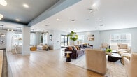 Located at 200–230 Pane Rd in Newington, Connecticut, this thoughtfully designed single-floor community amenity space offers a generous and versatile layout ideal for socializing and relaxation. The open floor plan is unified by light hardwood flooring and a neutral color palette of soft grays and whites, accented by distinctive steel-blue architectural details that lend the space a cohesive, modern character.

The main lounge area is anchored by a plush sectional sofa, comfortable swivel armchairs, and an area rug, all arranged to encourage conversation and comfort. Large indoor plants and curated artwork contribute warmth and visual interest throughout. A standout feature is the full-wall built-in media unit finished in muted blue-gray, complete with illuminated shelving, cabinetry, and a mounted flat-screen television.

A dedicated game area showcases a full-size shuffleboard table beneath stylish globe pendant lighting, with a tray ceiling detail painted in teal that defines the zone. Adjacent to this is a fully equipped kitchen and dining area with bar seating, making the space well-suited for hosted events and gatherings.

The entry corridor welcomes visitors with floor-to-ceiling glass double doors that flood the space with natural light and offer views to the outdoors. Additional seating nooks and open-concept zones throughout create a flexible, multi-use environment that balances both activity and quiet relaxation.