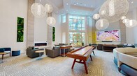 Located at 1231 Arya Way in Ocoee, Florida, this contemporary single-story amenity clubhouse showcases upscale design throughout its thoughtfully arranged communal spaces. The centerpiece is a sweeping open lounge area defined by dramatic double-height ceilings adorned with oversized spherical woven pendant lights that create a striking visual focal point. Floor-to-ceiling windows and glass doors flood the space with natural light while offering views of the landscaped outdoor surroundings. The lounge features multiple seating configurations with modern blue and gray upholstered furniture, a linear electric fireplace set within a wood-paneled accent wall, and vibrant vertical moss art installations that bring a natural, organic element indoors. A full-length shuffleboard table serves as a central game feature, complemented by a large-screen display for entertainment. A glass-wall-enclosed dining and kitchen area provides a dedicated space for gatherings, outfitted with dining tables, teal-accented chairs, and a fully equipped prep kitchen. A compact wet bar area with wood cabinetry, stone countertops, and a stainless steel refrigerator adds convenience for events. Decorative etched-glass double doors flanked by cylindrical columns provide elegant interior transitions, completing the polished and welcoming atmosphere of this community facility.