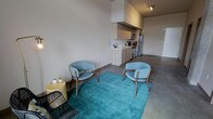 This single-floor space showcases a seamless open-plan layout that integrates the living area and kitchen into one cohesive environment. The polished concrete floors run throughout, lending an industrial-modern character that complements the neutral gray walls and clean architectural lines. Elevated ceilings add a sense of openness and volume to the space.

The living area is anchored by a vibrant teal area rug, which provides a bold pop of color against the understated palette. Two mid-century modern-style lounge chairs in soft blue upholstery with warm wood frames are arranged conversationally atop the rug, accompanied by a natural wood side table and a woven accent chair.

The kitchen sits neatly along the back wall, featuring warm-toned wood cabinetry that extends to the upper portion of the wall, creating a defined yet open cooking space. Stainless steel appliances—including a refrigerator, range, and microwave—are integrated seamlessly. A countertop sink and workspace complete the functional layout. Track lighting above the kitchen provides focused illumination. The overall ambiance is modern, understated, and thoughtfully styled.