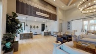 The Edison's leasing lounge and clubhouse is a thoughtfully designed single-story community facility that blends modern coastal aesthetics with functional, welcoming spaces. The main leasing lounge commands attention with its dramatic double-height ceilings adorned with a decorative scallop-patterned mural wall treatment, creating a striking visual centerpiece. Light-toned hardwood flooring runs throughout, complementing the neutral white and gray palette that defines the overall design language.

The leasing area features sleek white marble-topped reception desks accented with gray subway tile bases, paired with warm wood wishbone chairs and industrial-style pendant lighting. A dedicated workspace lined with windows allows ample natural light to fill the room, offering workstation seating along a wooden counter. Botanical and coastal-themed wall art adds character throughout.

A set of bold white sliding barn doors, labeled "Clubhouse," connects the leasing lounge to a separate lounge area appointed with upholstered seating, a stone fireplace surround, and additional gathering space. Scallop-patterned wallcovering, wall-mounted flat-screen televisions, and rustic wood console pieces add layered texture and functionality. Private offices with glass partition windows provide semi-transparent meeting spaces, ensuring both transparency and privacy within the facility.