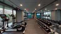 Located at 900 7th St SW in Washington, DC, this well-appointed fitness center offers a comprehensive workout environment designed with both functionality and aesthetics in mind. The space is organized into distinct workout zones, allowing users to move seamlessly between cardio, strength training, and functional fitness areas.

The cardio section is lined with Life Fitness treadmills, ellipticals, and a rowing machine, positioned along a wall of floor-to-ceiling windows that invite abundant natural light and provide views of the surrounding greenery — an energizing backdrop for any workout. Wall-mounted flat-screen televisions are strategically placed throughout to keep users entertained during their sessions.

The strength training area features a full rack of dumbbells, a cable and weight machine, and a dedicated space for functional training accessories including stability balls in multiple sizes. Warm wood-paneled walls with integrated shelving lend a refined, modern character to this section, complemented by large mirrors that visually expand the space.

The flooring throughout consists of durable rubber tiles, appropriate for the demands of a high-use fitness environment. Recessed lighting provides consistent, even illumination across the entire facility. A water fountain station and a small refreshment or equipment area with cabinetry are conveniently available, ensuring a complete and user-friendly fitness experience.