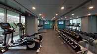 Located at 900 7th St SW in Washington, DC, this well-appointed fitness center offers a comprehensive workout environment designed with both functionality and aesthetics in mind. The space is organized into distinct workout zones, allowing users to move seamlessly between cardio, strength training, and functional fitness areas.

The cardio section is lined with Life Fitness treadmills, ellipticals, and a rowing machine, positioned along a wall of floor-to-ceiling windows that invite abundant natural light and provide views of the surrounding greenery — an energizing backdrop for any workout. Wall-mounted flat-screen televisions are strategically placed throughout to keep users entertained during their sessions.

The strength training area features a full rack of dumbbells, a cable and weight machine, and a dedicated space for functional training accessories including stability balls in multiple sizes. Warm wood-paneled walls with integrated shelving lend a refined, modern character to this section, complemented by large mirrors that visually expand the space.

The flooring throughout consists of durable rubber tiles, appropriate for the demands of a high-use fitness environment. Recessed lighting provides consistent, even illumination across the entire facility. A water fountain station and a small refreshment or equipment area with cabinetry are conveniently available, ensuring a complete and user-friendly fitness experience.