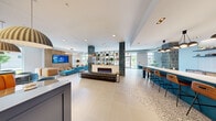 The common area at 1100 Broadway Street in Ann Arbor, Michigan is a thoughtfully designed, generously proportioned amenity space that blends contemporary style with functional versatility. The open-plan layout is anchored by a striking central lobby lounge featuring teal-glazed tile columns, a sleek linear fireplace set in a marble surround, and a built-in open shelving unit displayed between the columns. Large windows on multiple sides flood the space with abundant natural light and offer views of the surrounding streetscape.

The kitchen area is outfitted with a bold teal island topped with white quartz counters, bar-height seating, a herringbone-tiled backsplash, stainless steel appliances, and warm wood cabinetry — all illuminated by sculptural cluster pendant lighting. Adjacent terrazzo flooring transitions seamlessly between zones, tying the design together with a polished yet approachable aesthetic.

A dedicated lounge zone features plush teal velvet sofas, orange banquette booths, and a vibrant artistic feature wall, creating an inviting social atmosphere. A separate conference room provides a focused meeting environment with a long wood table, cognac leather chairs, patterned wallcovering, large windows, and a wall-mounted display screen. The space as a whole reflects a vibrant, design-forward character ideal for community gathering and collaboration.