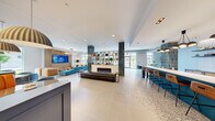 The common area at 1100 Broadway Street in Ann Arbor, Michigan is a thoughtfully designed, generously proportioned amenity space that blends contemporary style with functional versatility. The open-plan layout is anchored by a striking central lobby lounge featuring teal-glazed tile columns, a sleek linear fireplace set in a marble surround, and a built-in open shelving unit displayed between the columns. Large windows on multiple sides flood the space with abundant natural light and offer views of the surrounding streetscape.

The kitchen area is outfitted with a bold teal island topped with white quartz counters, bar-height seating, a herringbone-tiled backsplash, stainless steel appliances, and warm wood cabinetry — all illuminated by sculptural cluster pendant lighting. Adjacent terrazzo flooring transitions seamlessly between zones, tying the design together with a polished yet approachable aesthetic.

A dedicated lounge zone features plush teal velvet sofas, orange banquette booths, and a vibrant artistic feature wall, creating an inviting social atmosphere. A separate conference room provides a focused meeting environment with a long wood table, cognac leather chairs, patterned wallcovering, large windows, and a wall-mounted display screen. The space as a whole reflects a vibrant, design-forward character ideal for community gathering and collaboration.