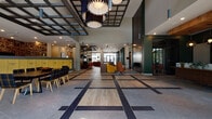 This single-story amenity space showcases a thoughtfully designed interior that balances contemporary style with functional comfort. The expansive lobby serves as the heart of the space, distinguished by a striking geometric floor pattern that blends light wood planks with dark tile inlays, creating a dynamic visual foundation underfoot. Soaring ceilings are punctuated by an industrial-modern grid framework from which sculptural pendant lights hang, casting warm, layered illumination across the open floor plan.

The lounge area features a rich cognac leather sectional sofa, a circular area rug, and a wall-mounted television, offering a relaxed gathering spot. Nearby, a green diamond-grid bookshelf adds a pop of color and a cozy, literary touch. A teal-tiled fireplace accent wall anchors one corridor, adding depth and warmth. The mailroom area showcases a bright yellow bank of package lockers, while a separate café-style service counter with teal cabinetry, dark countertops, and bold geometric wallcovering provides a welcoming refreshment station with outdoor views.

The fitness center is generously equipped with cardio machines, strength-training equipment, and a standout bouldering climbing wall set against an exposed brick backdrop. A checkered carpet in green and grey tones ties the athletic space together, while large windows invite natural light throughout.