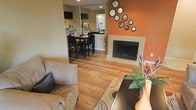 This single-floor residence offers a thoughtfully designed open-concept layout that seamlessly connects the living, dining, and kitchen areas, creating a cohesive and functional living environment. The living room is anchored by a striking fireplace set against a bold terracotta-orange accent wall, which adds warmth and visual interest to the space. A decorative arrangement of circular convex mirrors adorns the accent wall, lending an artistic touch to the room. Rich hardwood flooring flows throughout, tying the spaces together with a consistent, warm aesthetic. The living area is generously sized, accommodating comfortable seating and a central area rug that defines the space. Adjacent to the living room, the dining area features a dark-toned table and chair set, ideal for everyday meals and entertaining. Beyond the dining space, the kitchen is visible through an open pass-through, showcasing dark countertops and ample cabinetry. Recessed lighting throughout provides clean, even illumination across all areas. The overall ambiance of the home is welcoming and well-appointed, blending contemporary design elements with warm, earthy tones to create an inviting atmosphere throughout.