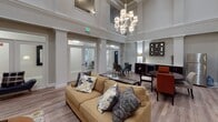 This single-floor lounge space is defined by its striking architectural presence, most notably its soaring double-height ceilings adorned with detailed crown molding and a statement chandelier that anchors the main living area. The open layout is finished with warm wood-look flooring throughout, lending a cohesive and inviting character to the entire space.

The lounge area serves as the centerpiece, furnished with a generous tan sofa and an upholstered round ottoman atop a patterned area rug. A tiled fireplace surround rises prominently along one wall, topped with a wall-mounted flat-screen television, while a pair of gray armchairs creates a cozy conversation nook nearby.

Adjacent to the lounge, a dining area features a round dark-wood table paired with distinctive orange upholstered chairs, complemented by a dark credenza and decorative wall mirrors. A kitchenette along the wall provides a stainless steel refrigerator, a beverage cooler, and ample cabinetry with light countertops.

Frosted glass-paneled doors connect to adjoining rooms, enhancing the sense of openness. The neutral warm-gray palette, layered lighting, and carefully curated décor combine to create an atmosphere that is both polished and welcoming.
