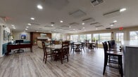 Located at 5001 SW 20th St in Ocala, Florida, this single-story community clubhouse offers a well-appointed gathering space designed to serve residents with a range of amenities. The welcoming entryway features warm wood-look flooring, teal wainscoting accents, and decorative framed artwork showcasing the surrounding community, setting an inviting tone throughout.

The expansive main common area seamlessly integrates a dining section and a lounge area beneath a ceiling fitted with recessed lighting and geometric acoustic panels. Multiple round dining tables with wood chairs provide generous seating, while large windows along the rear wall flood the space with natural light and offer views of an outdoor pool and patio area. A striking full-height brick fireplace anchors the lounge side of the room, accompanied by comfortable sofas and accent chairs arranged on colorful area rugs.

The modern kitchen area is separated by a sleek glass partition wall, featuring contemporary dark cabinetry, light countertops, stainless steel appliances, and a central island — ideal for community events and gatherings. An adjacent office space includes a workstation and framed property photographs, serving administrative needs. Consistent hardwood-style flooring and the teal-and-beige color palette tie the entire facility together into a cohesive, functional, and welcoming environment.