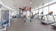 This spacious fitness center is thoughtfully designed to support a full range of workout disciplines within a bright, modern environment. The space is defined by expansive floor-to-ceiling windows that line the perimeter, flooding the interior with natural light and offering open outdoor views, including skyline and palm tree scenery that enhances the overall workout experience.

The flooring throughout consists of durable, textured grey fitness tiles that provide both traction and comfort. Industrial-style pendant and drum lights hang from elevated white ceilings, contributing to a clean, contemporary aesthetic. Large mirrors along the walls visually expand the space and support form monitoring during training.

The cardio zone features multiple treadmills, an elliptical trainer, and a stationary bike, all positioned to take advantage of the panoramic views. A central multi-functional rig with an orange steel frame serves as the focal point of the strength area, accommodating pull-ups, suspension training, and functional fitness movements. A TITLE-branded heavy punching bag hangs from the rig, supporting boxing and conditioning training. Additional strength equipment includes cable machines, adjustable benches, a dumbbell rack, plate-loaded machines, and a dedicated ab station, making this fitness center a comprehensive training facility.