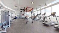 This spacious fitness center is thoughtfully designed to support a full range of workout disciplines within a bright, modern environment. The space is defined by expansive floor-to-ceiling windows that line the perimeter, flooding the interior with natural light and offering open outdoor views, including skyline and palm tree scenery that enhances the overall workout experience.

The flooring throughout consists of durable, textured grey fitness tiles that provide both traction and comfort. Industrial-style pendant and drum lights hang from elevated white ceilings, contributing to a clean, contemporary aesthetic. Large mirrors along the walls visually expand the space and support form monitoring during training.

The cardio zone features multiple treadmills, an elliptical trainer, and a stationary bike, all positioned to take advantage of the panoramic views. A central multi-functional rig with an orange steel frame serves as the focal point of the strength area, accommodating pull-ups, suspension training, and functional fitness movements. A TITLE-branded heavy punching bag hangs from the rig, supporting boxing and conditioning training. Additional strength equipment includes cable machines, adjustable benches, a dumbbell rack, plate-loaded machines, and a dedicated ab station, making this fitness center a comprehensive training facility.