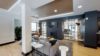 This single-floor space presents a refined and carefully curated lounge environment defined by a striking dark tile feature wall that anchors the room. A built-in fireplace sits centrally within the feature wall beneath a wall-mounted flat-screen television, creating a natural focal point. The ceiling incorporates a warm wood-paneled tray detail that adds architectural depth and contrasts elegantly with the white structural columns and surrounding walls.

The seating area is furnished with plush grey sofas and accent chairs arranged atop a textured area rug, complemented by a mix of marble-topped side tables and mirrored accent pieces. Gold-framed open display shelving units flank the feature wall on both sides, adding a glamorous, contemporary touch while providing display and storage opportunities.

Natural light enters through large windows along the perimeter, brightening the hardwood flooring that runs throughout. A deep teal accent wall adds a bold color contrast to the otherwise neutral palette. The overall ambiance is polished and modern, balancing warm materials with sleek, reflective finishes to create an inviting yet sophisticated atmosphere.