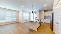 This single-floor residence offers a thoughtfully designed open-concept layout with two bedrooms and two full bathrooms. Light-toned wood flooring runs consistently throughout the space, creating a cohesive and airy feel across all rooms.

The kitchen serves as a central feature, anchored by a spacious island with a dark quartz countertop, an undermount sink with a matte black faucet, and generous seating potential. White shaker-style cabinetry lines the walls, complemented by a tile backsplash, stainless steel appliances including a refrigerator, range, and microwave, and black hardware accents. Dual pendant lights hang above the island, adding a modern touch. Double doors nearby provide ample pantry or storage space.

The open living and dining area flows directly from the kitchen, bathed in natural light from multiple windows fitted with horizontal blinds. Recessed lighting supplements the natural light throughout.

The primary bedroom is generously sized and features a ceiling fan, while the en-suite bathroom includes a single vanity with quartz countertops, white cabinetry with black hardware, and access to a walk-in closet. The second bathroom mirrors this refined aesthetic with a vanity, tub-shower combination, and matching finishes.