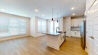 This single-floor residence offers a thoughtfully designed open-concept layout with two bedrooms and two full bathrooms. Light-toned wood flooring runs consistently throughout the space, creating a cohesive and airy feel across all rooms.

The kitchen serves as a central feature, anchored by a spacious island with a dark quartz countertop, an undermount sink with a matte black faucet, and generous seating potential. White shaker-style cabinetry lines the walls, complemented by a tile backsplash, stainless steel appliances including a refrigerator, range, and microwave, and black hardware accents. Dual pendant lights hang above the island, adding a modern touch. Double doors nearby provide ample pantry or storage space.

The open living and dining area flows directly from the kitchen, bathed in natural light from multiple windows fitted with horizontal blinds. Recessed lighting supplements the natural light throughout.

The primary bedroom is generously sized and features a ceiling fan, while the en-suite bathroom includes a single vanity with quartz countertops, white cabinetry with black hardware, and access to a walk-in closet. The second bathroom mirrors this refined aesthetic with a vanity, tub-shower combination, and matching finishes.