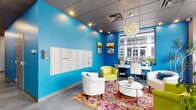 Located at 408 S Nevada Ave in Colorado Springs, Colorado, this single-story lobby space makes a striking first impression through its bold and contemporary design. The interior is defined by vivid turquoise accent walls that contrast sharply with neutral gray surfaces, creating an energetic and welcoming atmosphere. Polished concrete flooring runs throughout, lending an urban, modern character to the space.

Natural light floods the interior through expansive floor-to-ceiling windows along the street-facing facade, offering clear views of the surrounding Colorado Springs streetscape and connecting the interior to the outdoors. A sculptural, globe-shaped chandelier serves as a standout decorative focal point overhead, complementing the drop tile ceiling fitted with recessed lighting.

The lounge area is furnished with a curated mix of white and chartreuse accent chairs arranged around a circular white coffee table, anchored by a vibrant multicolored area rug that adds depth and personality to the space. A wall-mounted bank of individual mailboxes is integrated seamlessly into the blue accent wall, indicating a functional role within a larger building. A dedicated desk area with shelving is positioned near the windows, providing a practical workspace. Potted greenery and colorful artwork complete the polished, lively ambiance of this well-appointed lobby.