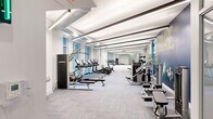 Located at 1100 Broadway Street in Ann Arbor, Michigan, this modern fitness center offers a comprehensive workout environment spread across a generously sized single-floor layout. The space is divided into two distinct functional zones, each designed to support a different style of training.

The main gym floor features a clean, contemporary aesthetic with patterned carpet tiles, exposed ductwork set against a bright white ceiling, and linear LED lighting that runs the length of the room. Large windows along one wall flood the space with natural light and provide views of the surrounding greenery. This primary zone is lined with Life Fitness cardio equipment—including treadmills, ellipticals, rowing machines, and stationary bikes—alongside a full complement of strength machines, cable systems, free weights, barbells, and benches. Full-length mirrors along the far wall enhance the sense of depth. A bold motivational mural reading "The Stronger You Are, The Better You Feel" anchors the strength training area, adding energy to the environment.

A secondary studio space features light hardwood-style flooring and accommodates functional and high-intensity training, with equipment including spin bikes, a punching bag, medicine balls, kettlebells, TRX suspension trainers, and a multi-functional training rig. A water fountain station and climate control are also available on-site for user convenience.