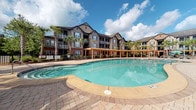 Located at 1475 Benton Blvd in Pooler, Georgia, this outdoor amenity space is designed to deliver a resort-like experience within a residential community setting. At its centerpiece is a generously sized swimming pool with a smooth, curvilinear shape and clear turquoise water, surrounded by a broad paver deck finished in warm-toned brick-pattern stone. Lounge chairs are arranged poolside, offering ample space for sunbathing beneath the Georgia sky.

Anchoring the patio area are multiple substantial wood pergolas with open-beam construction, providing partial shade over seating areas and walkways. The pergolas frame the space architecturally and lend a natural, warm aesthetic that complements the surrounding brick and neutral-toned residential buildings visible in the background. Tall palm trees are interspersed throughout, reinforcing the tropical, leisure-focused atmosphere.

A standout feature of the outdoor space is the built-in outdoor kitchen, which includes a stainless steel grill set into a stacked-stone counter wall — ideal for outdoor gatherings. Nearby, umbrella-shaded high-top bistro tables and chairs provide casual dining and socializing areas. The entire patio is finished in interlocking pavers, creating a cohesive and polished look throughout the space.